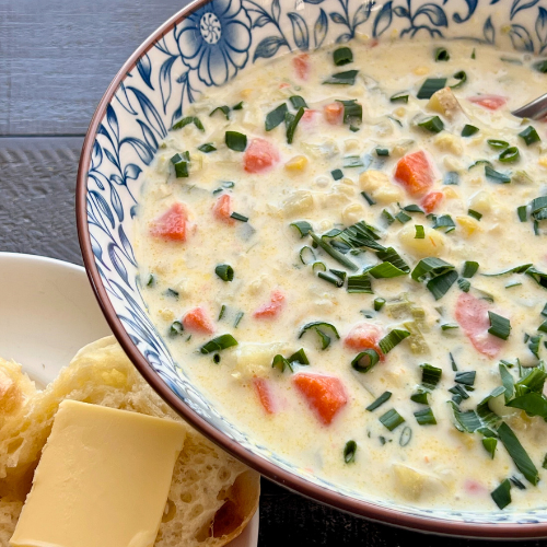 A large bowl of fresh corn chowder garnished with green onions sits on a countertop next to a roll with butter.