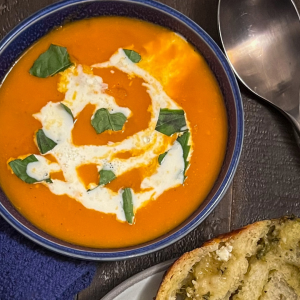 A blue bowl with red roasted garlic, tomato, and white bean creamy soup topped with a swirl of cream and served with a slice of pesto babka